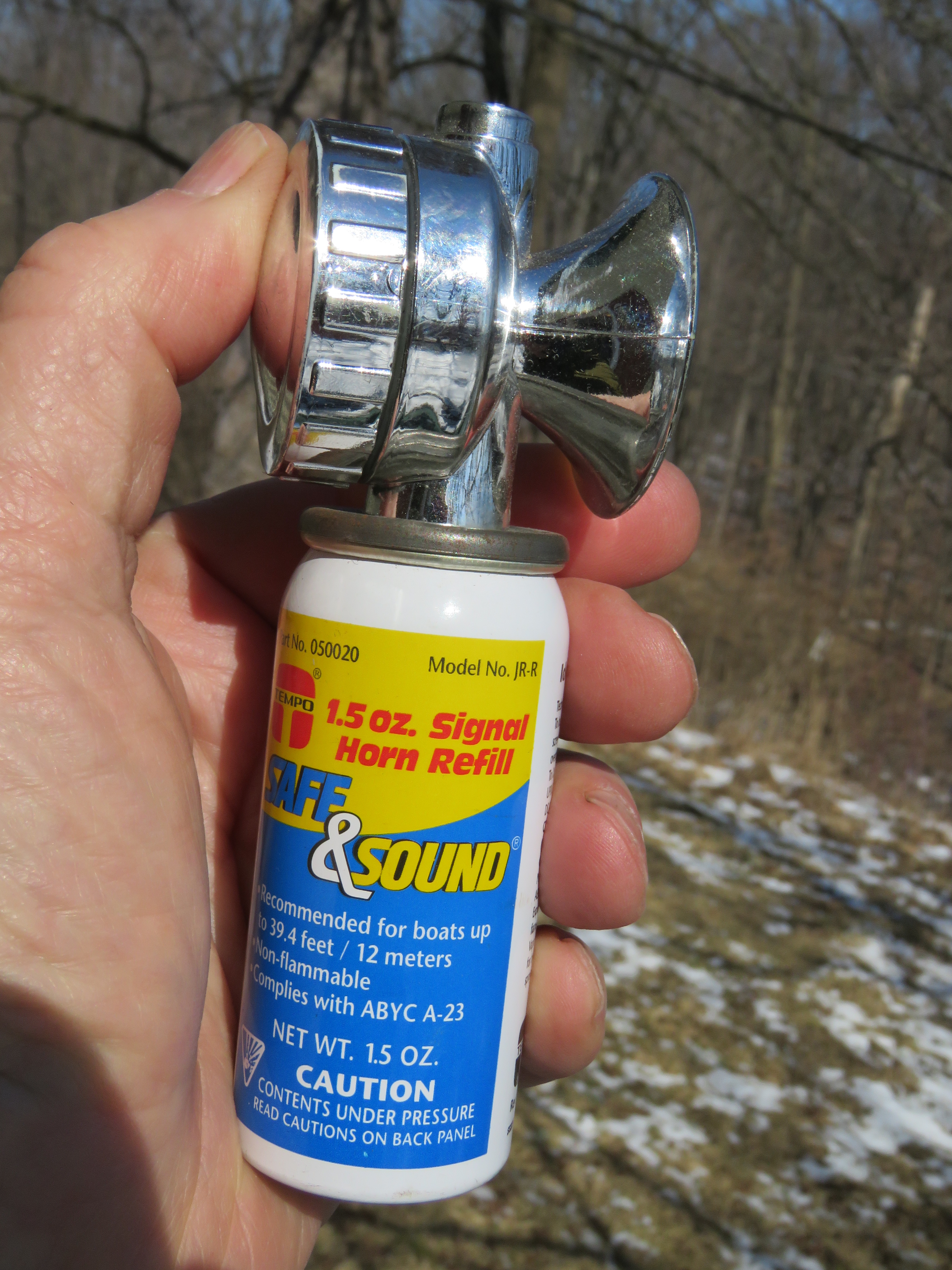 A person holding a 1.5 oz signal horn refill canister with a chrome horn nozzle in a natural setting, featuring trees and a snowy ground.
