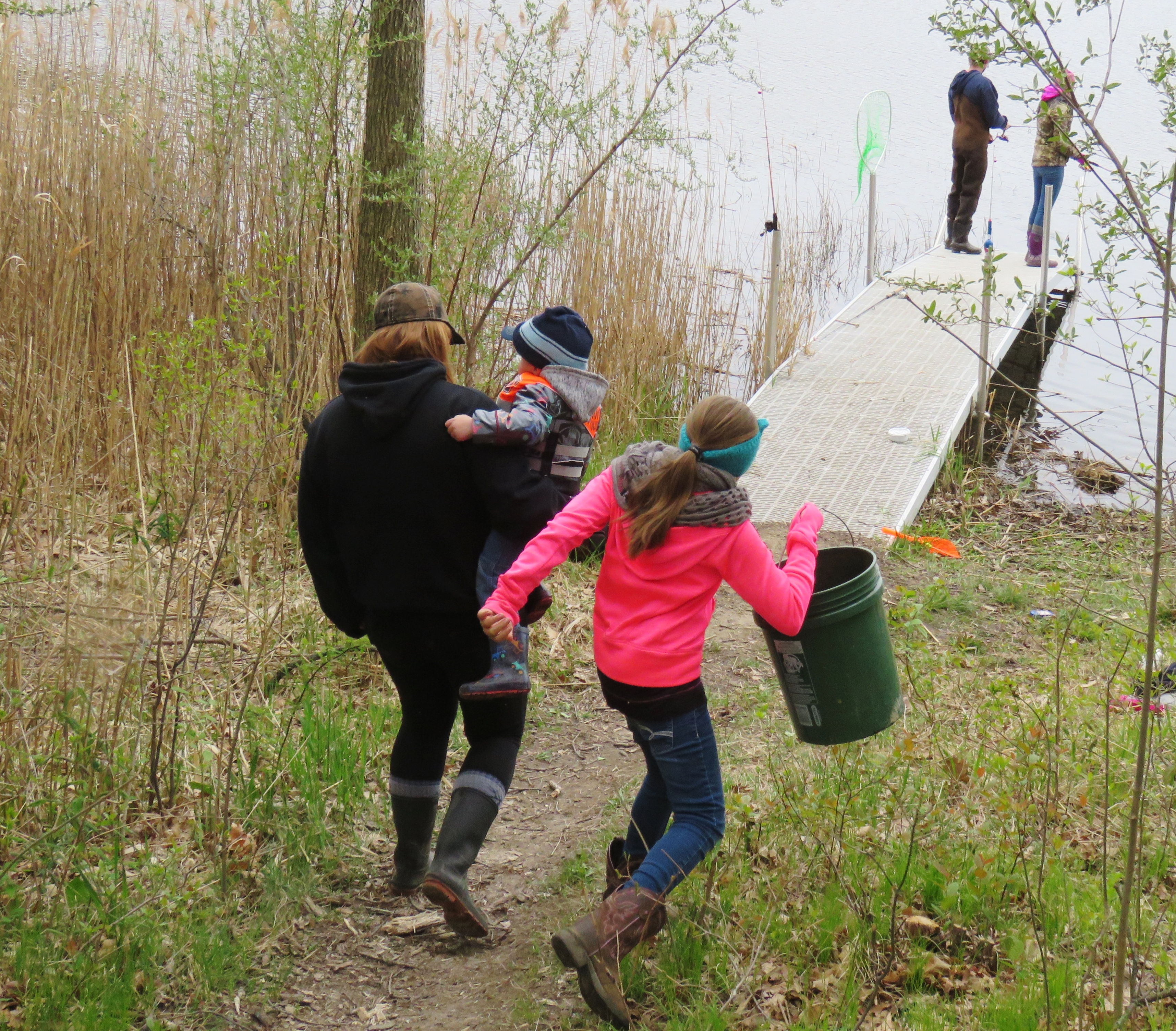 A woman carrying a young child while walking along a path by the water, accompanied by another girl holding a bucket. In the background, a dock extends into the water with two people fishing.
