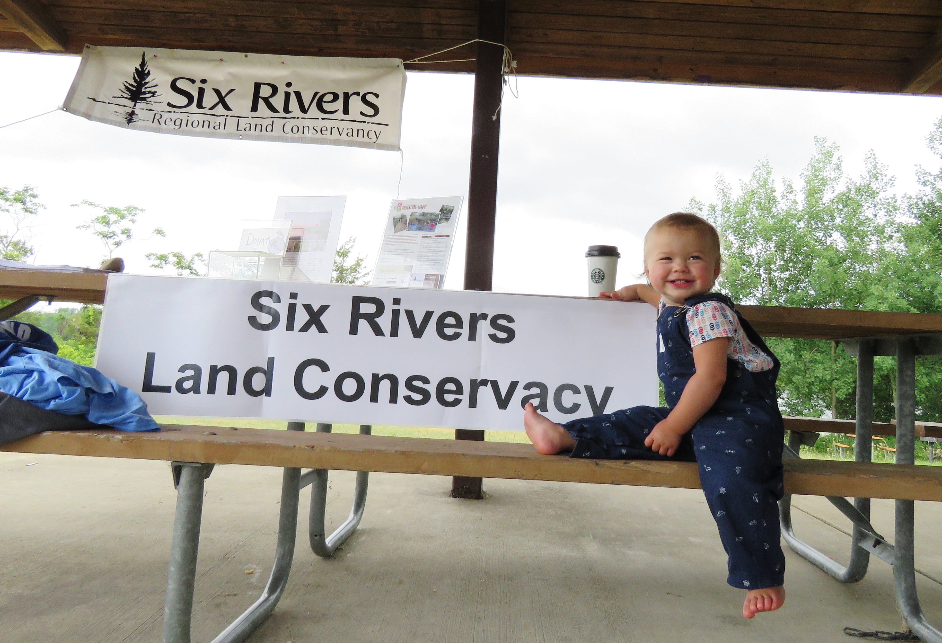 A happy toddler sitting on a picnic table with a 'Six Rivers Land Conservancy' banner in the background.