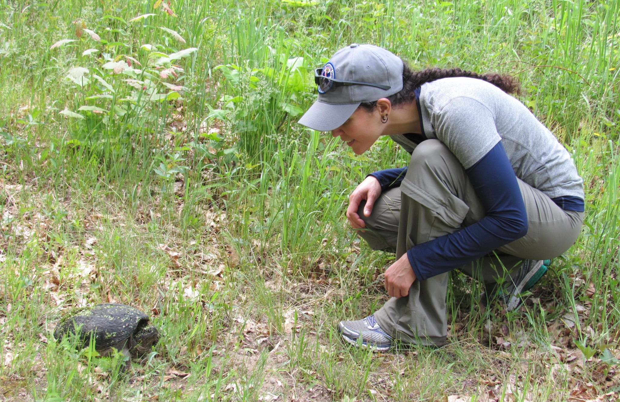 A person squatting on the ground, observing a turtle in a grassy area surrounded by plants and foliage.