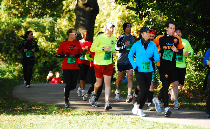 A group of runners jog down a paved path on a sunny fall day. The leaves on the trees at the edge of the trail are mostly green, just starting to change to fall colors.