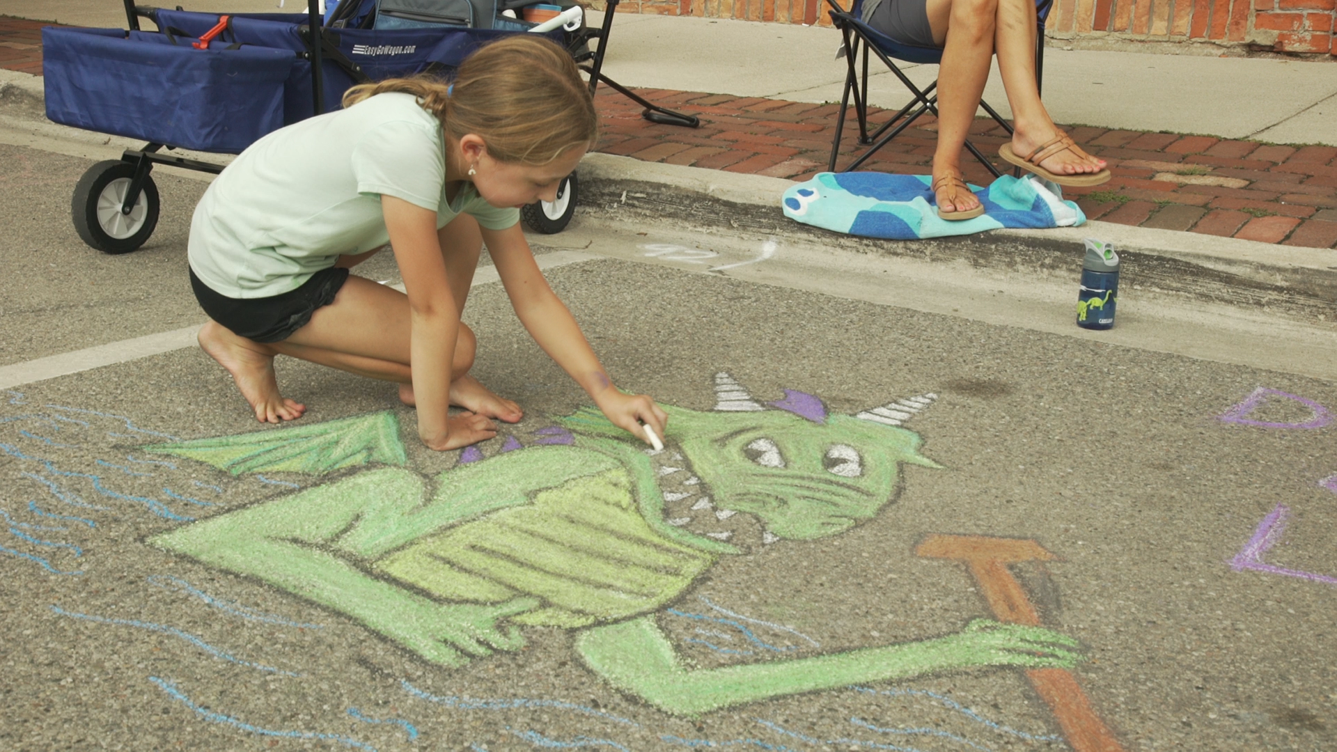 A young girl crouches down as she draws a dragon in chalk during Lake Orion's Dragon on the Lake Chalk Art Challenge. 