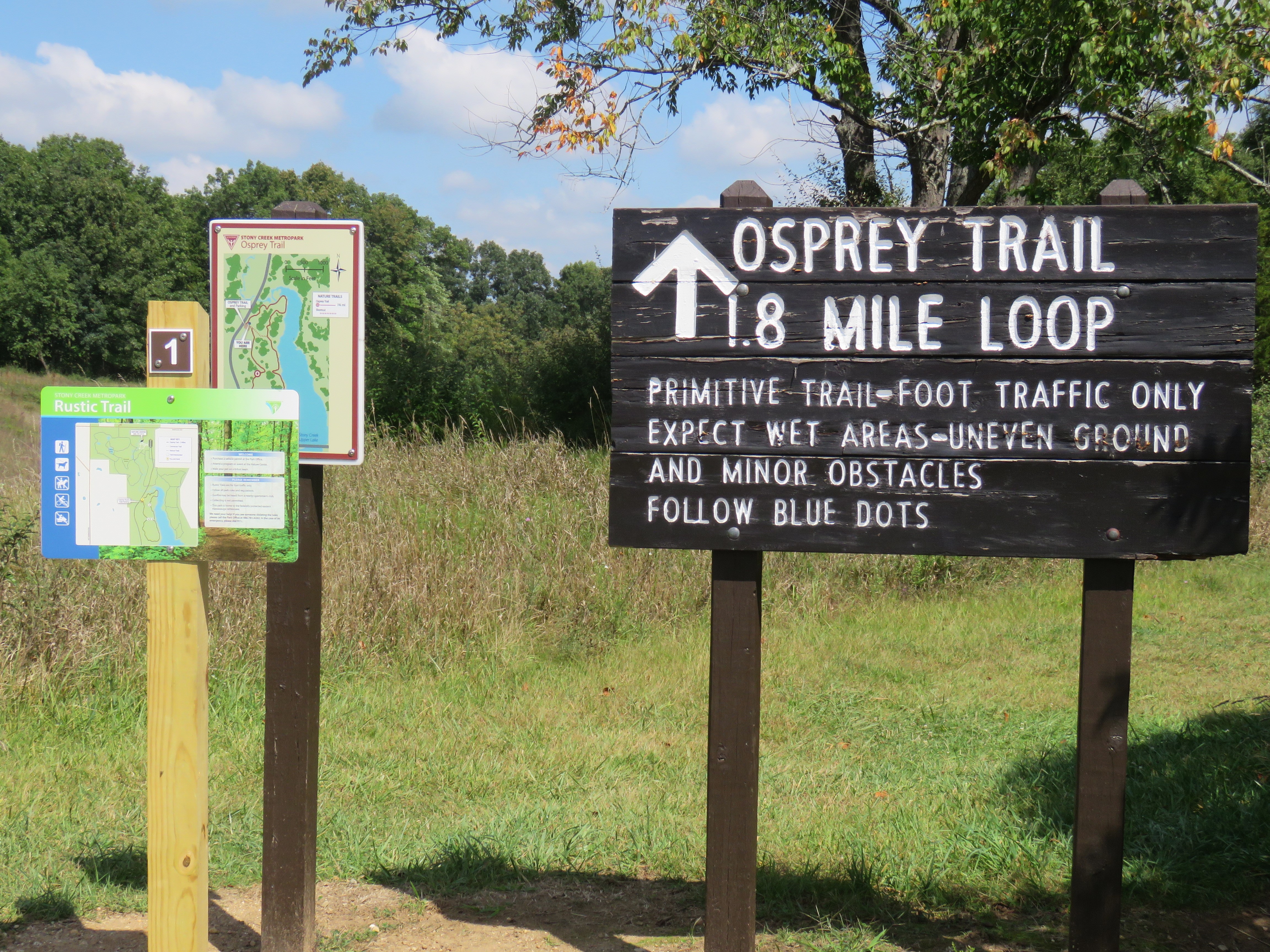 A photograph of three interpretive signs and their wooden posts in Stony Creek Metropark. Two smaller signs to the left show maps. A large dark brown wooden sign on the right shows an arrow pointing up. The text reads: Osprey Trail 1.8. Mile Loop. Underneath in a smaller font, it reads: Primitive Trail-Foot Traffic Only Expect Wet Areas-Uneven Ground and Minor Obstacles Follow Blue Dots