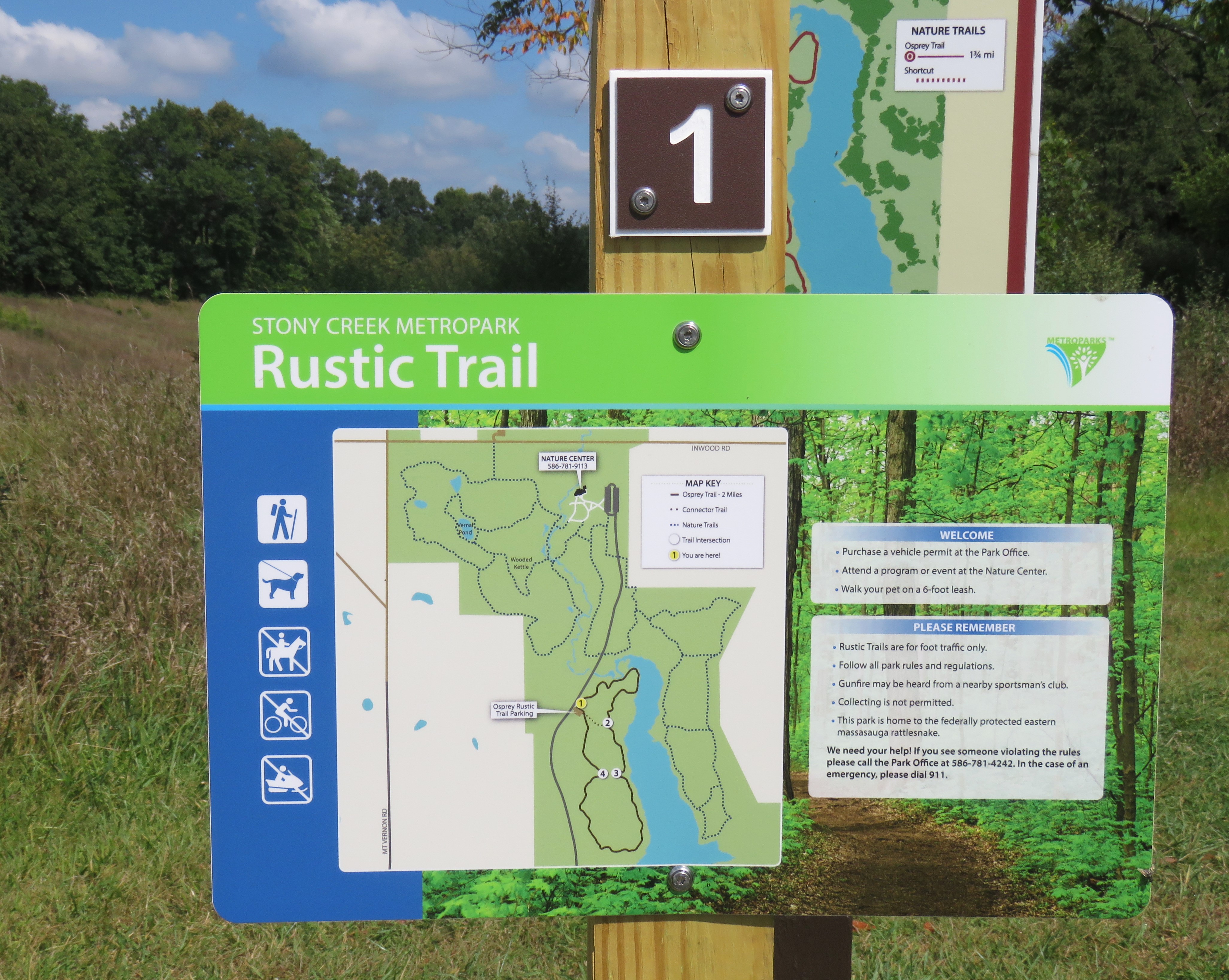 A close up of an interpretive sign at Stony Creek Metropark. It is titled, Rustic Trail and shows a map and some text with the headings "Welcome" and "Please Remember." Trees can be seen in the distance and the sky above is blue with white puffy clouds. 