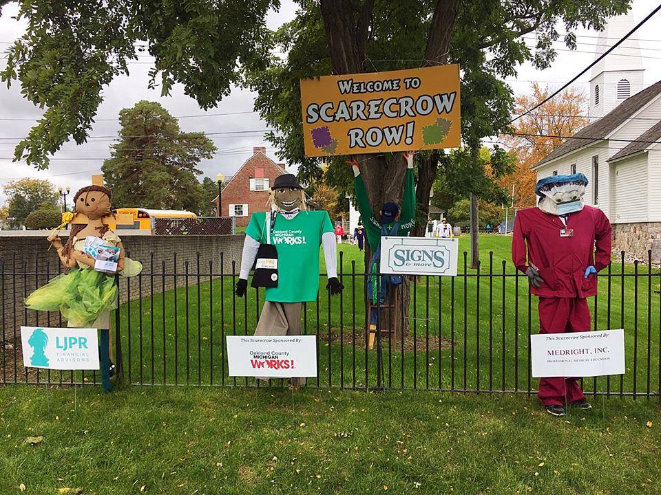 Three scarecrows lined up along a black wrought-iron fence. Signs in front show the businesses that decorated them. A sign hanging from a large maple tree behind the scarecrows reads: Welcome to Scarecrow Row!