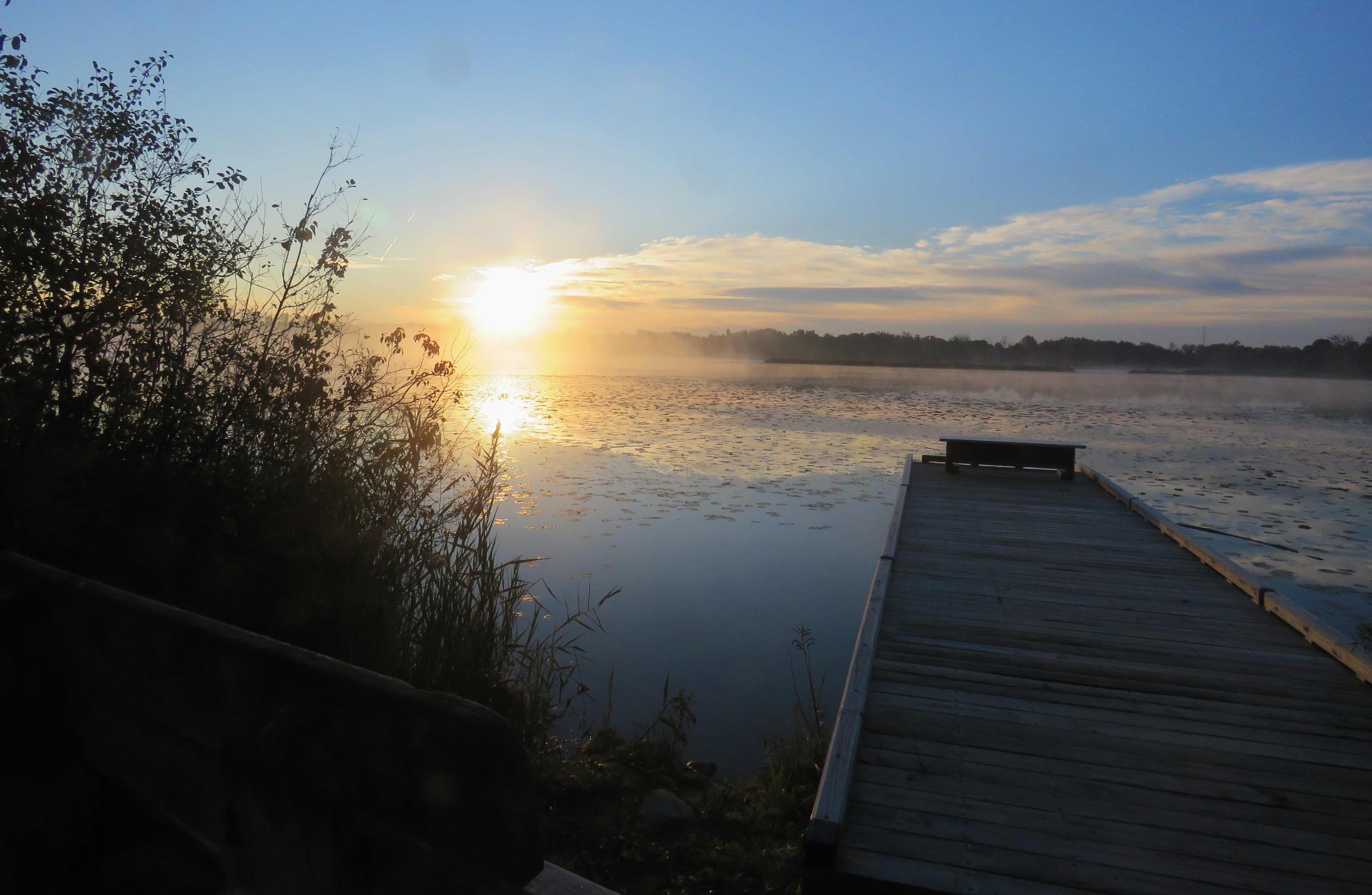 The sun rises over Lake Sixteen in Lake Orion. A portion of a wooden dock stretches from land and out over the lake. Fog rises up over the lake. 