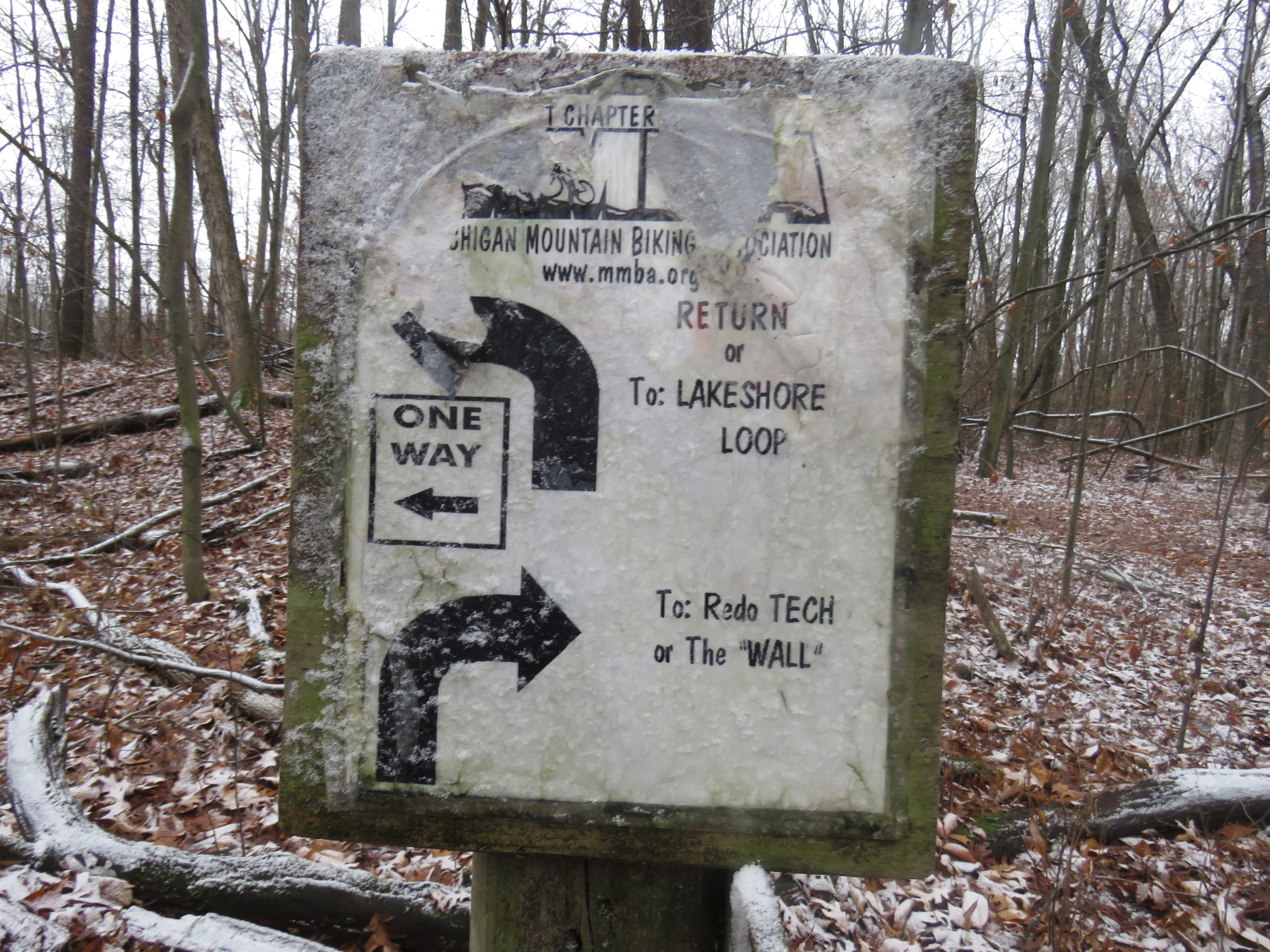 A trail marker with a ragged sign that has arrows pointing to RETURN or Lakeshore Loop, and Redo TECH or The "Wall."