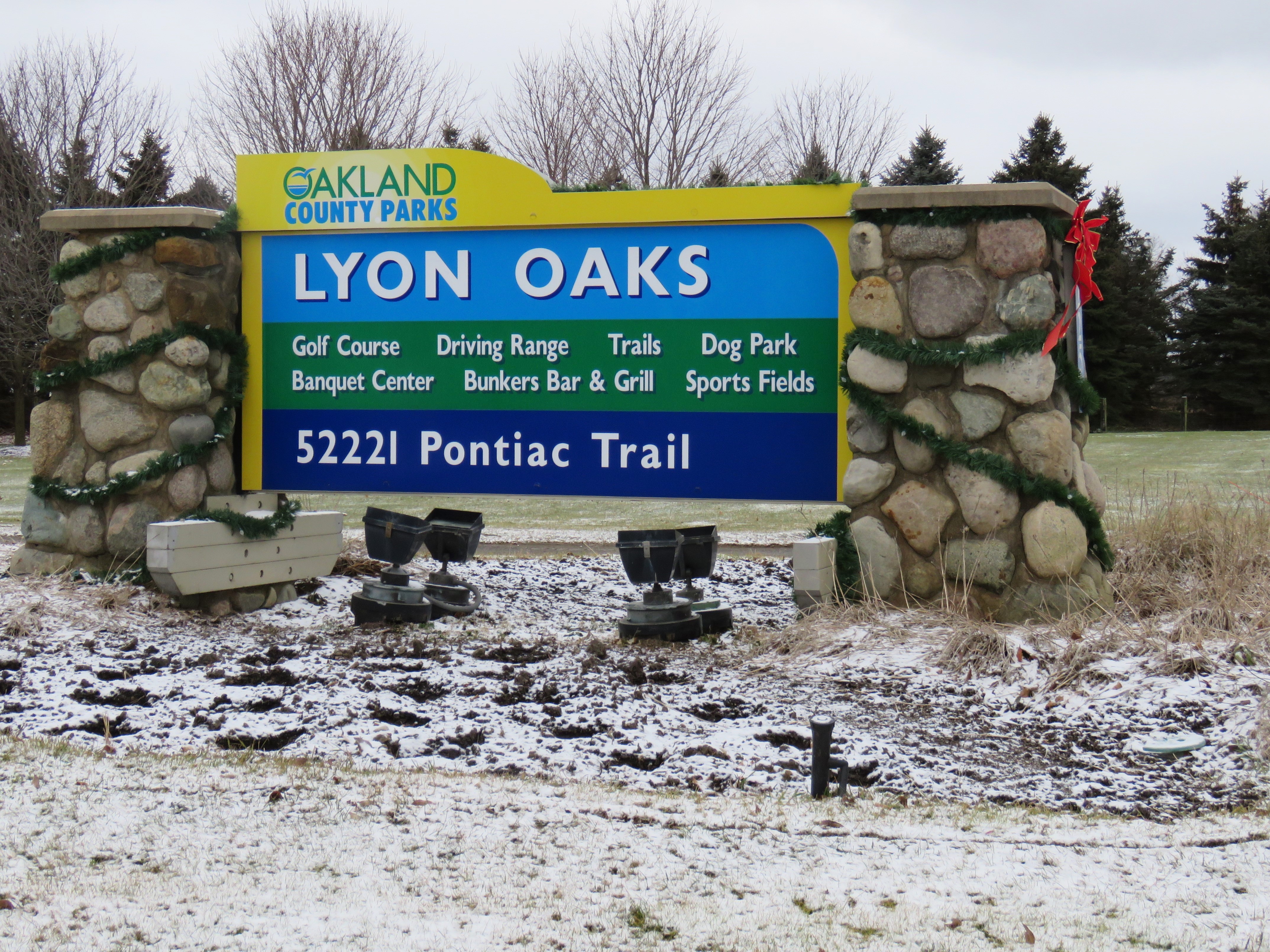 A large sign reads: Oakland County Parks, Lyon Oaks 52221 Pontiac Trail. Amenities listed on the sign are: Golf Course, Banquet Center, Driving Ranet, Bunkers Bar & Grill, Trails, Dog Park, and Sports Fields