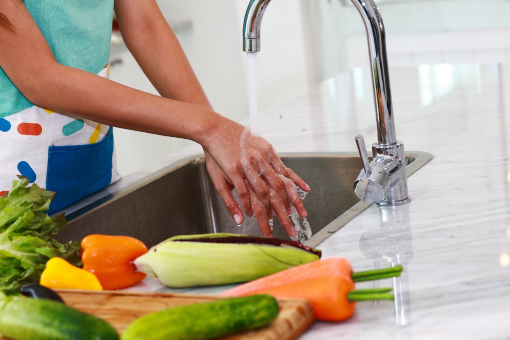 Young woman washing hands in the kitchen sink with veggies on a cutting board next to her.